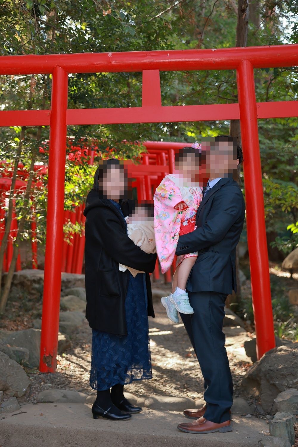 大宮氷川神社の朱色の鳥居の下で、紺のワンピースの母親が赤ちゃんを、スーツの父親が着物の娘を抱いているところ(お顔は伏せています)